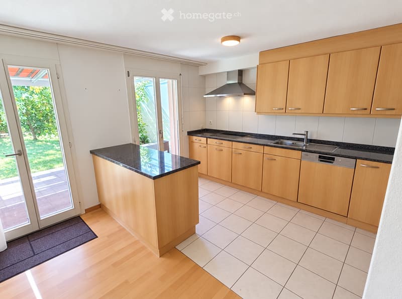 Modern kitchen with light wooden cabinets, black countertops, a central island, and built-in appliances including a dishwasher and range hood. The space features large windows and a sliding glass door leading to an outdoor area.