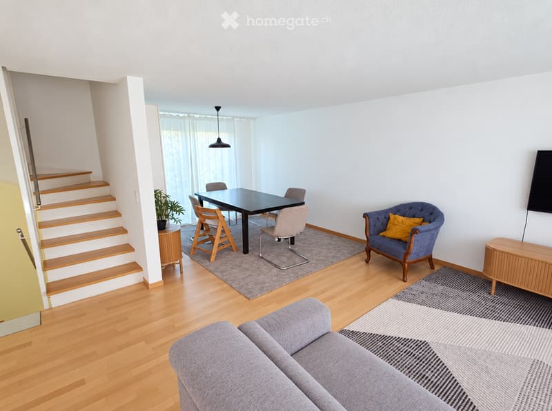 Bright, modern living room with wooden flooring, a grey sofa, a blue armchair with a yellow pillow, a black dining table with four chairs, a small wooden side table with a plant, and a television on a wooden stand.