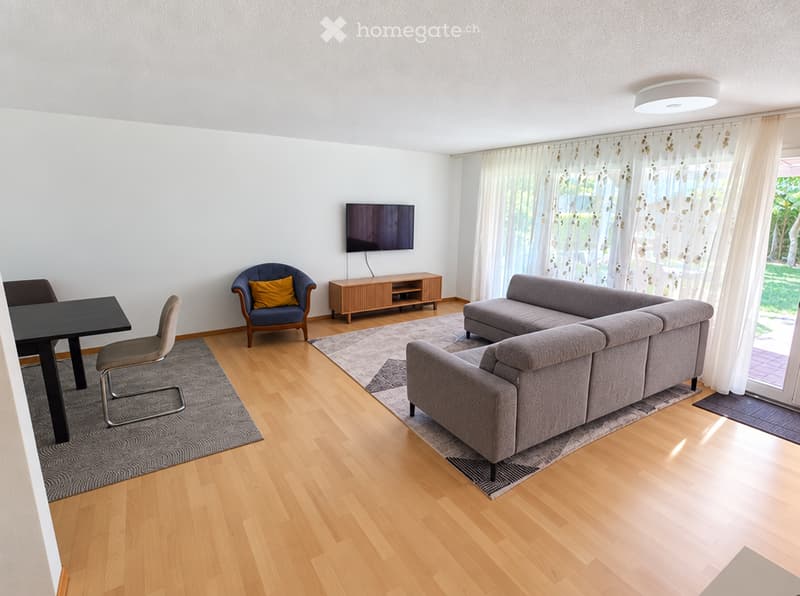 Modern living room with light wooden flooring, grey sectional sofa, blue armchair, black dining table, and a flat-screen TV on a wooden stand.