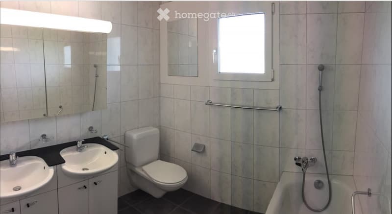 Modern bathroom with beige tiled walls, a double sink vanity, a toilet, a bathtub with a handheld shower, and a large mirror with built-in lighting.