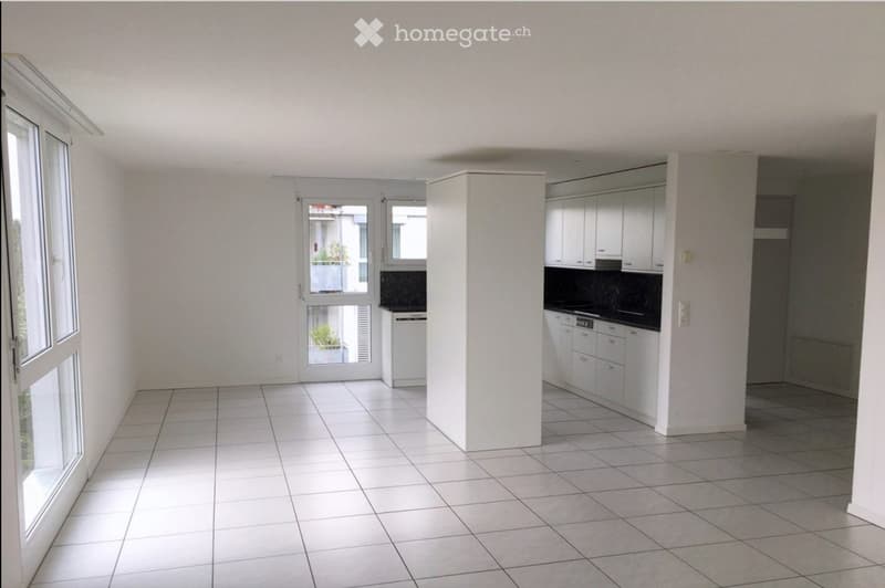 Empty room with white tiled flooring, white walls, and large windows. The space includes a small kitchen area with white cabinets and a countertop.