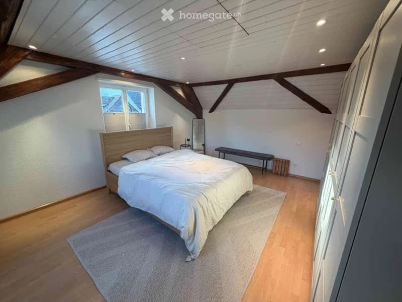 Cozy attic bedroom with wooden beams, white walls, and wooden flooring. Features a large bed with white bedding, a bench, a nightstand, and a mirror.