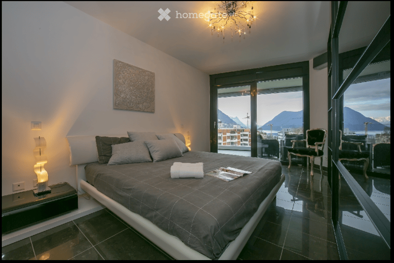 Spacious bedroom with a large bed, gray bedding, and a view of the mountains. A chandelier and a lamp are on the wall.