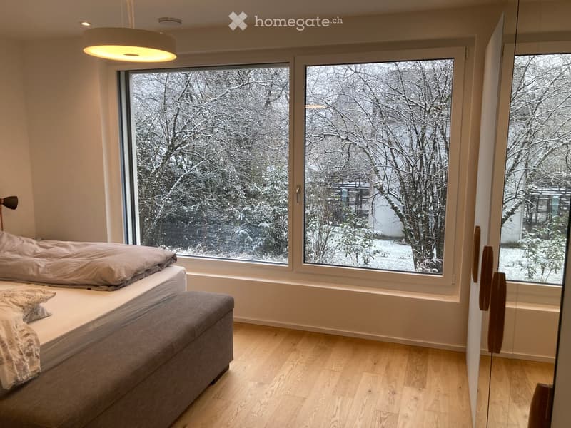 Bright bedroom with large windows overlooking a snowy outdoor scene, featuring a bed with a grey duvet and an ottoman.