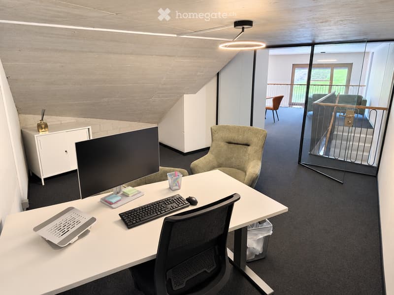 Modern office space with a white desk, ergonomic chair, laptop, keyboard, mouse, and screen. A small cabinet with a decorative vase is visible, and a comfortable armchair is placed nearby. The room features exposed concrete walls and a sleek, contemporary