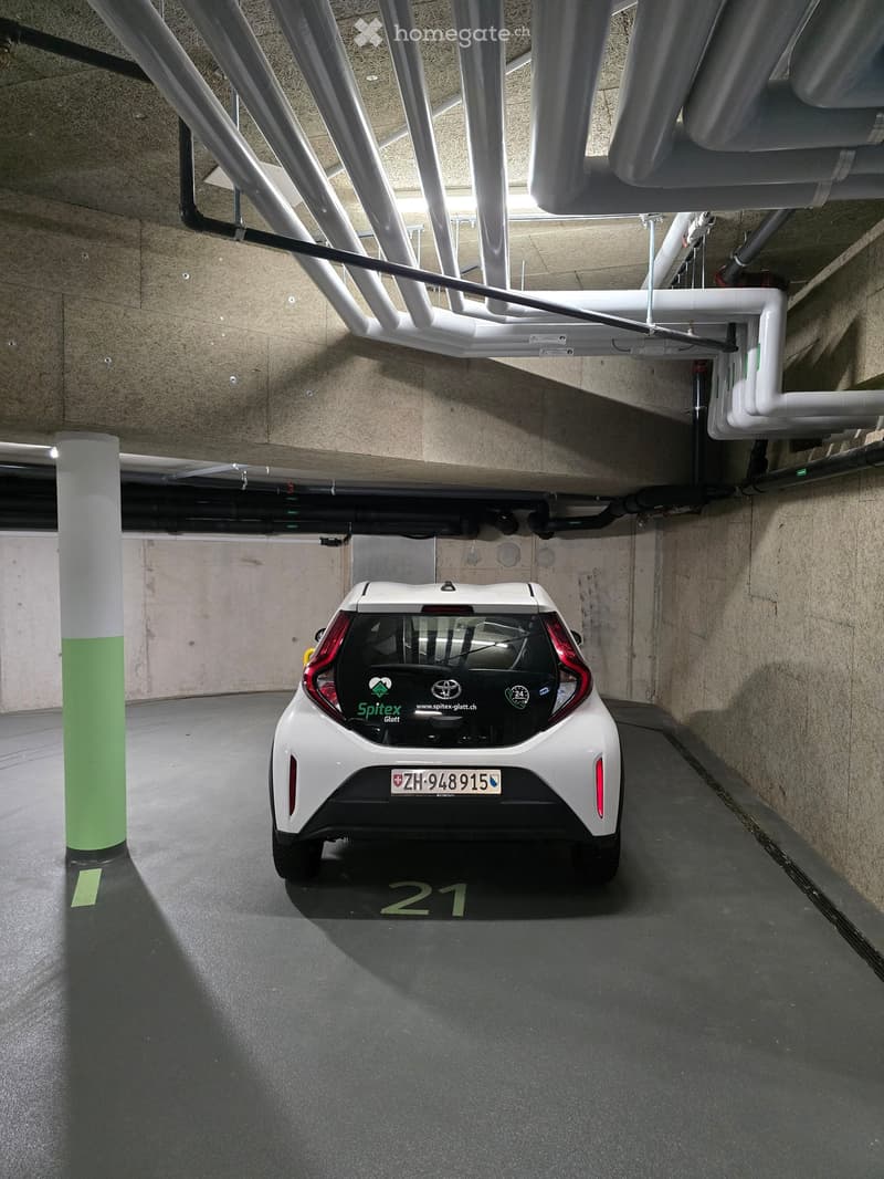 Underground parking area with a white car parked in bay number 21, surrounded by concrete pillars and exposed pipes.