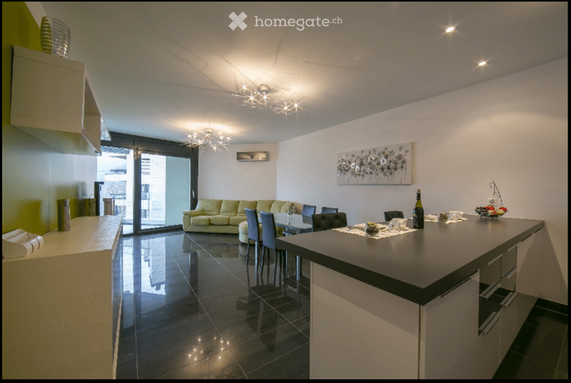Open-plan living and dining area with a modern sofa, dining table with chairs, kitchen island, artwork on the wall, and a bottle on the counter.