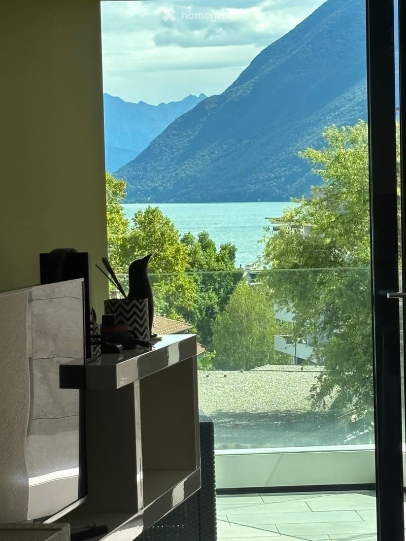 Large window with view of mountains and lake, desk with decorative items, glass door opening to terrace
