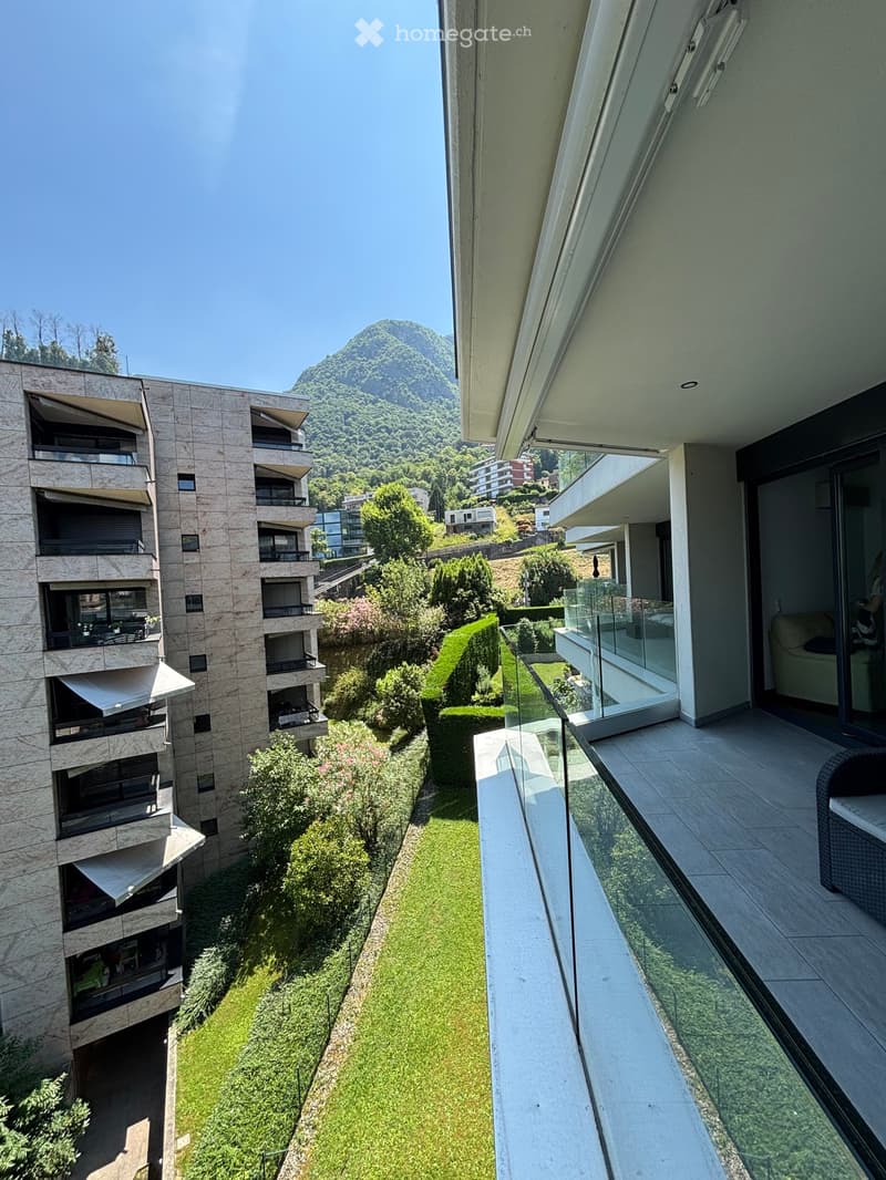 Modern apartment with a balcony overlooking a green area and distant mountains. It has a glass railing, a tiled floor, and a couch inside.