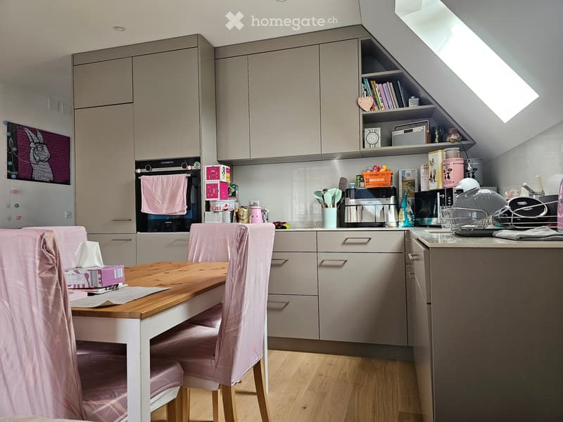 Modern kitchen with grey cabinets, wooden dining table with pink chairs, various appliances including oven, microwave, and fridge, and shelves with books and decorative items.