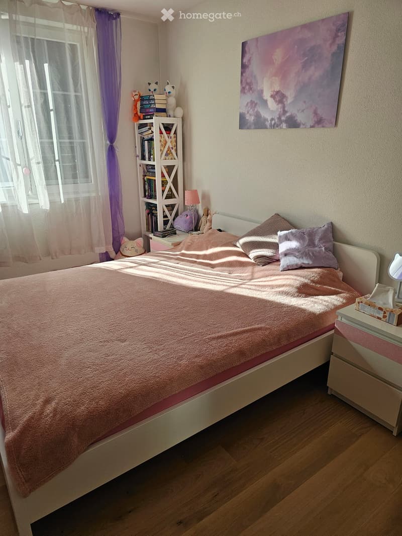 Cozy bedroom with a white bed, pink bedding, nightstand, bookshelf, and decorative items. Natural light streams in through a window with sheer curtains.