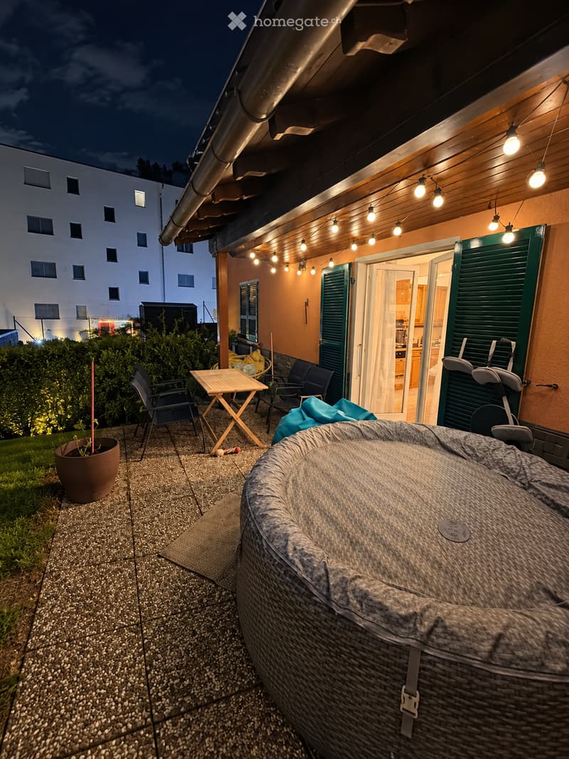 Outdoor patio area with a hot tub, small table, chairs, and a potted plant, illuminated by string lights.