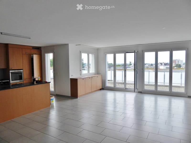 Modern kitchen with light wooden cabinets, stainless steel oven, and large windows leading to a balcony.