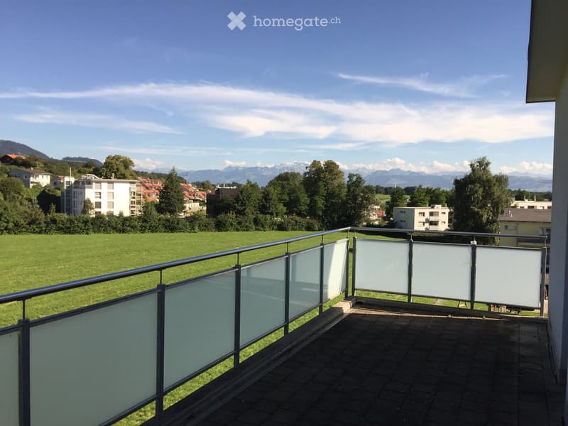Balcony with glass balustrade overlooking a green field and distant mountains.