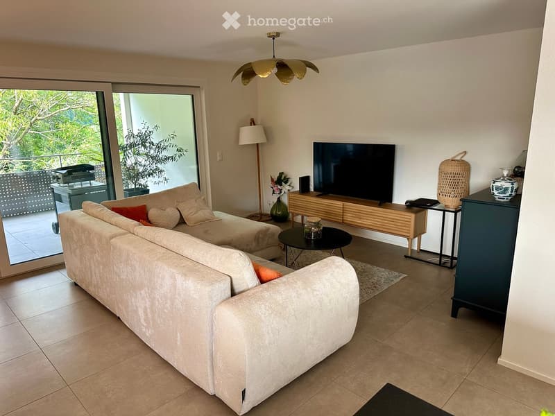 Modern living room with a large sofa, coffee table, television, floor lamp, side table, and decorative items. Large sliding glass doors open to a balcony with outdoor furniture.