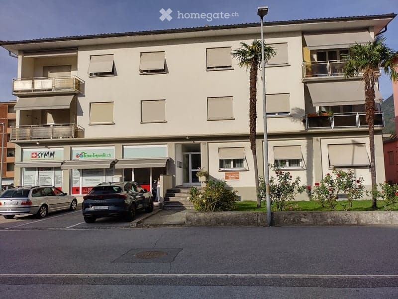 Three-story building with a modern facade, multiple windows with shutters, and a small entrance area. There are two cars parked in front, and the building has a small garden area with palm trees and shrubs.
