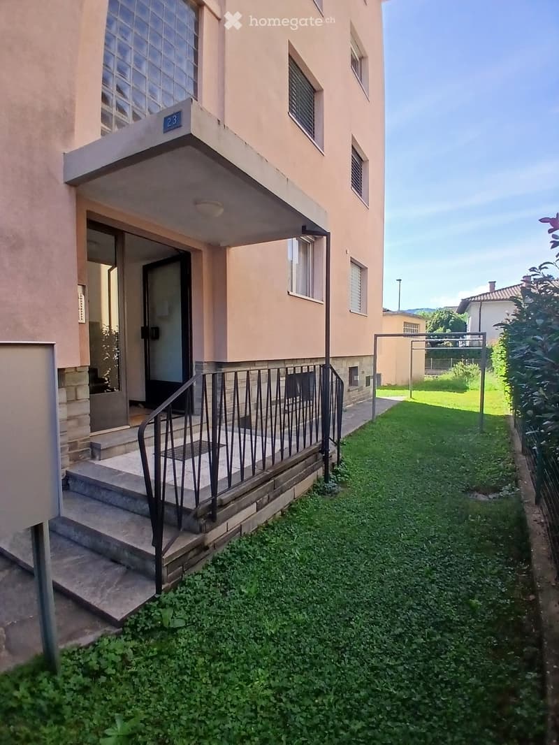 Two-story residential building with a covered entrance, steps, and a small garden area with grass and plants.