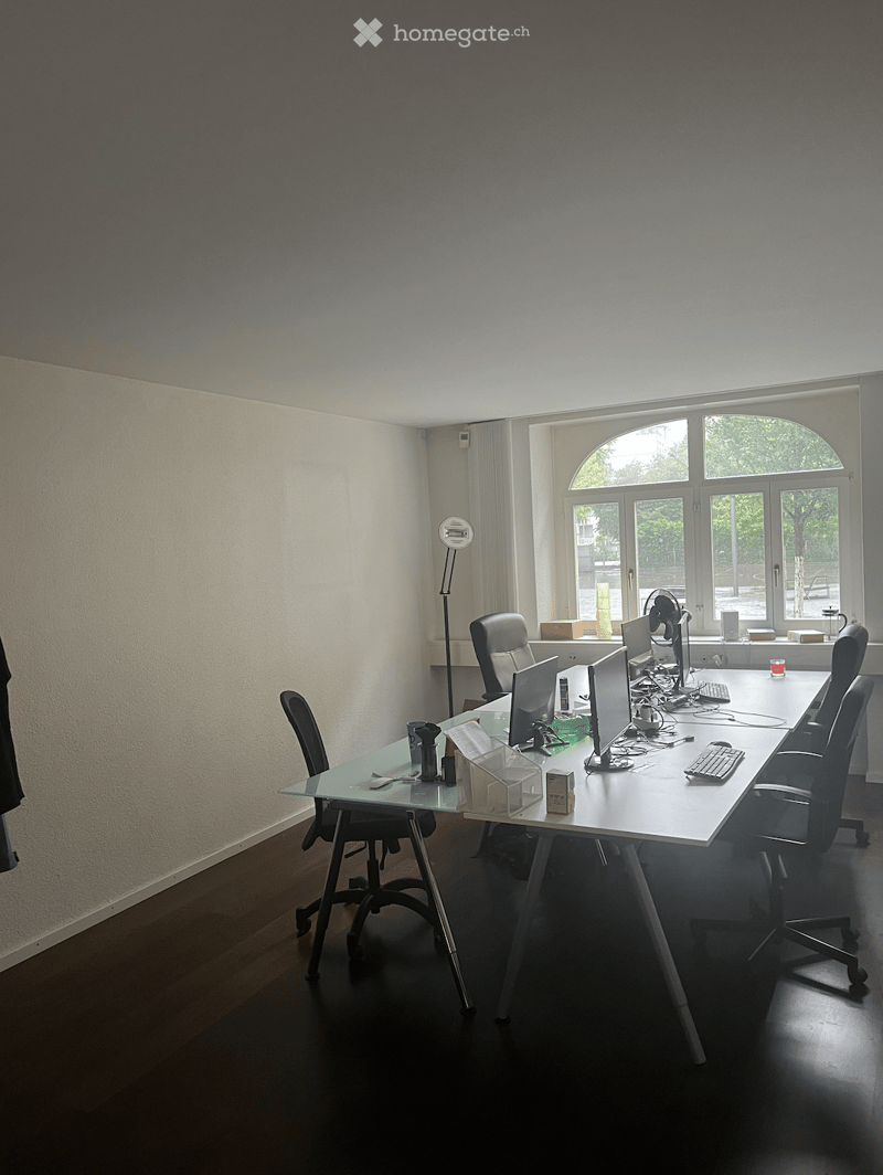 Office room, 6 desk chairs, long desk with computers, monitors, keyboards, and mouses, white walls, lamp, office supplies on the desk, windows.