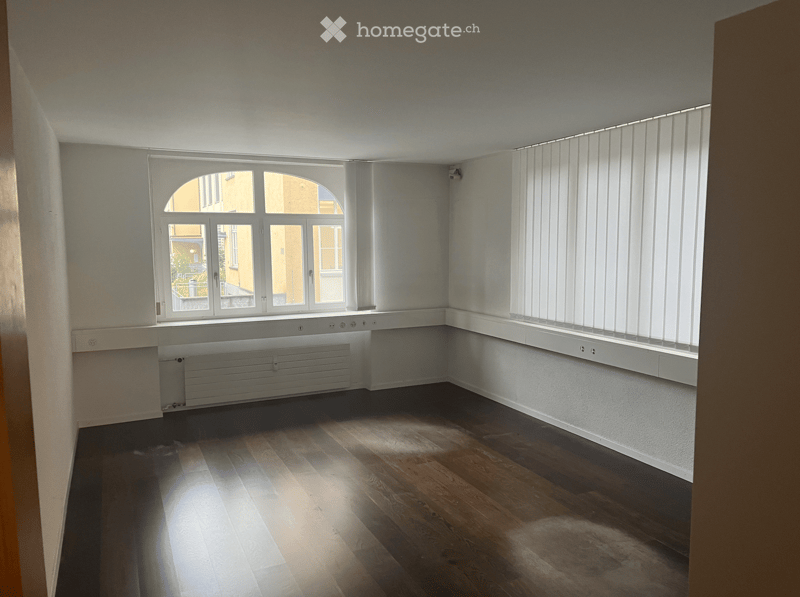 empty room, white walls, wooden floor, two windows with blinds, small heating system