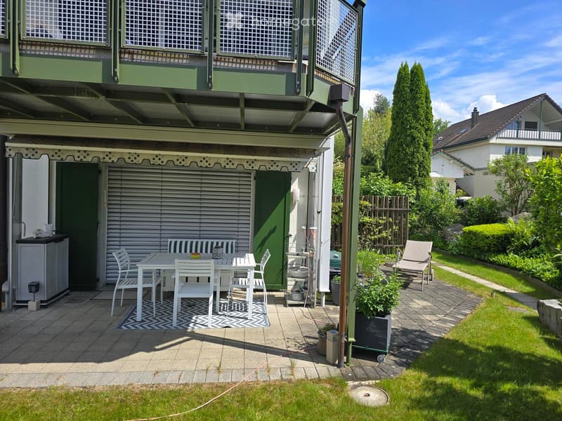 Outdoor patio with a table, chairs, bench, and umbrella, surrounded by a well-maintained garden and lawn.