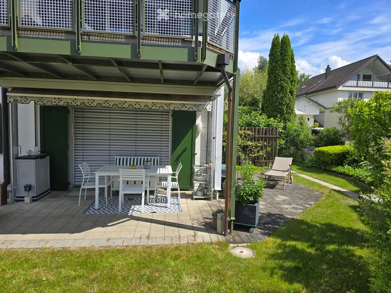 Garden with a patio area featuring a table, chairs, and a bench, surrounded by greenery and a well-maintained lawn.
