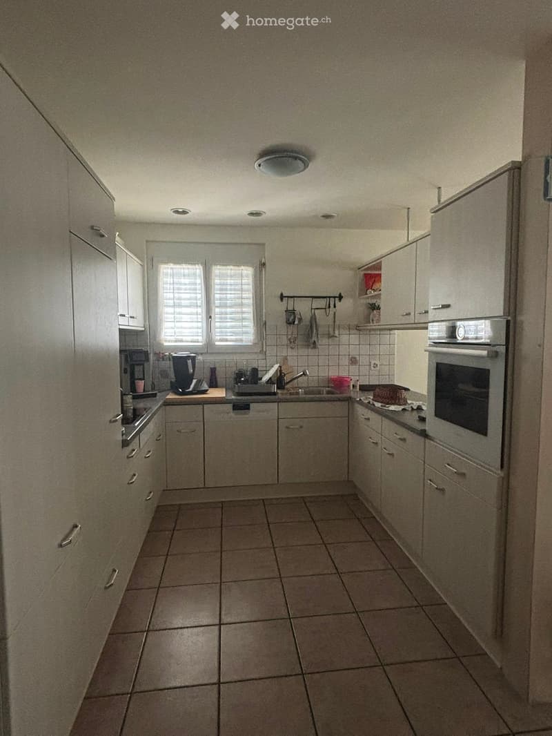 A modern kitchen with light-colored cabinets, tiled floor, and stainless steel appliances including an oven, sink, and dishwasher. The space features a window with blinds and a countertop with various items.