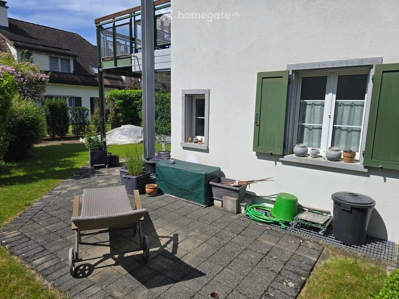 Garden patio area with a covered bench, potted plants, a wheelbarrow, and a hose reel. The house has white walls, green shutters, and a balcony with decorative items on the windowsill.