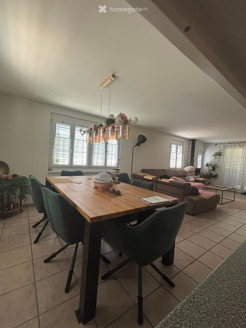 A modern living and dining area with a large wooden dining table, green chairs, a brown sofa, and a hanging chandelier with floral decorations.