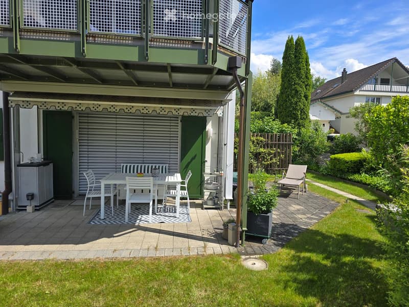 Garden area with a patio, white table and chairs, a small sofa, and a lounge chair, surrounded by greenery and a well-maintained lawn.