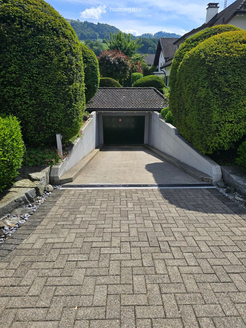 Driveway leading to a garage with a tiled roof, surrounded by trimmed hedges and trees, with a house visible in the background.