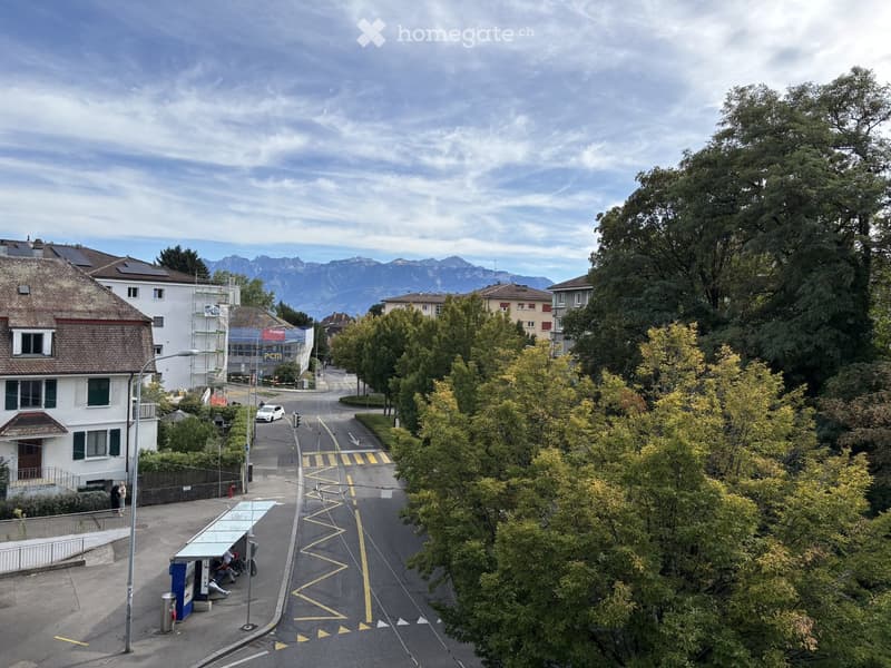 A quiet residential street lined with trees and houses, with a distant view of mountains. A bus stop is visible on the left, and a few pedestrians are walking along the sidewalk.