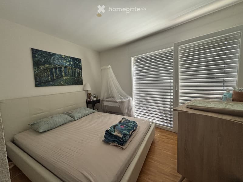 A well-lit bedroom with a large bed, pillows, a nightstand with a lamp, a mirror, and a painting on the wall. The room features wooden flooring and large windows with blinds.