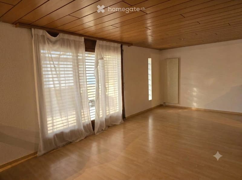 Empty room with wooden flooring, white walls, large window with blinds, and a radiator on one wall.