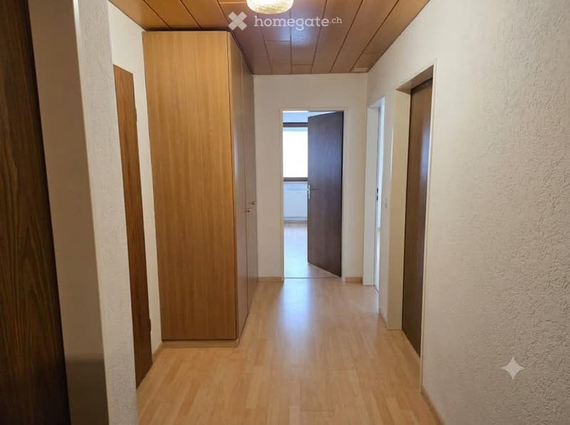 A clean, well-lit hallway with wooden flooring, white walls, and a wooden cabinet on the left side. The hallway leads to an open doorway at the end.