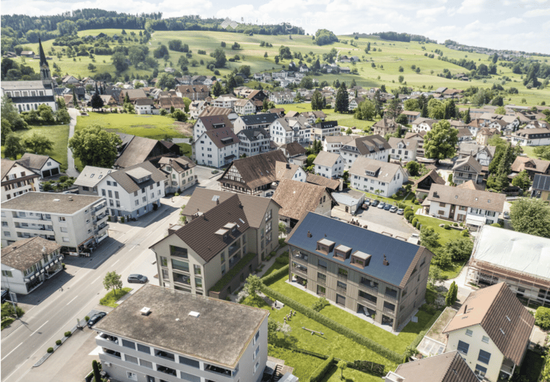 Aerial view of a small town with a mix of residential and commercial buildings, surrounded by green fields and hills.