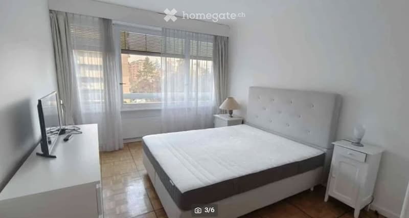 Modern bedroom with a large bed, nightstands, a lamp, and a TV screen on a stand. The room has large windows with curtains and a tiled floor.