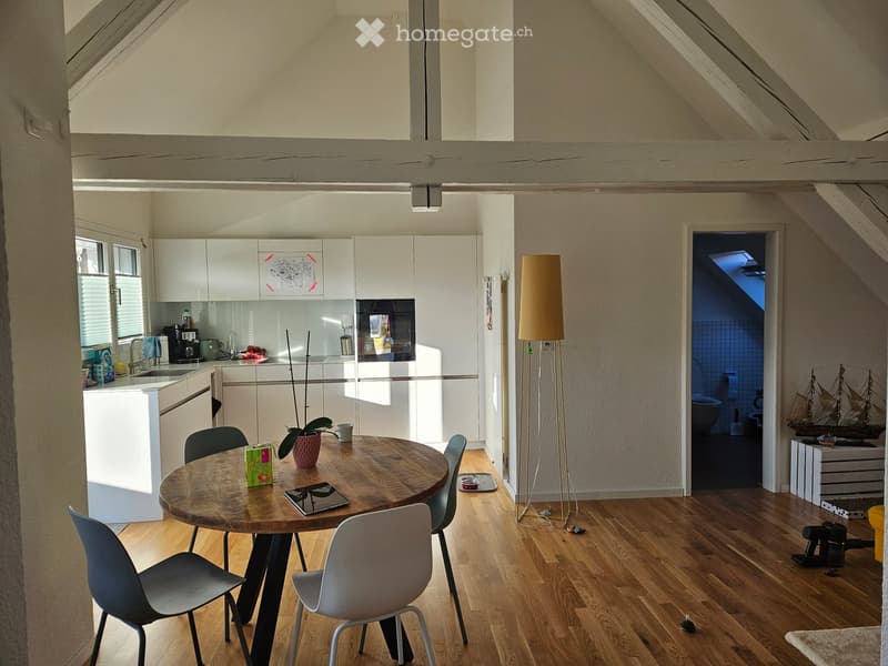 Bright, open-plan attic space with wooden floors, white cabinets, a round wooden dining table with chairs, a floor lamp, and a small kitchen area with a sink and refrigerator.