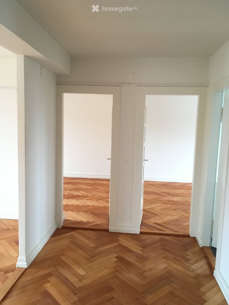 Empty hallway with wooden herringbone flooring and white walls, featuring two open doorways leading to other rooms.