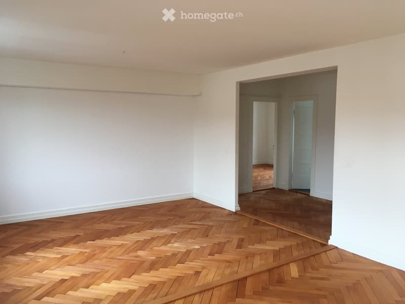 Empty room with wooden herringbone flooring and white walls, featuring an open doorway leading to another room.