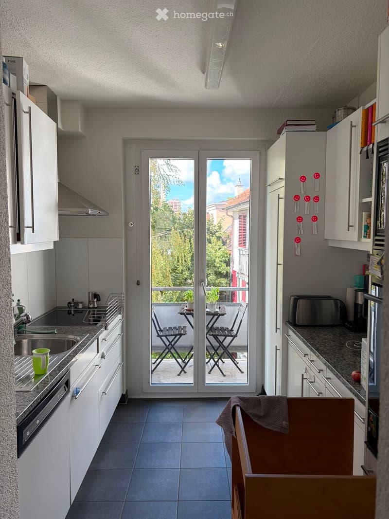 Modern kitchen with white cabinets, granite countertops, stainless steel appliances, and a sliding glass door leading to a small balcony with a table and chairs.