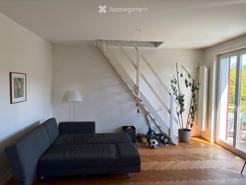 Living room with a grey sectional sofa, wooden floor, white walls, a staircase leading to an upper level, a large potted plant near the sliding glass door, and a floor lamp. A picture frame hangs on the wall, and toys are scattered near the staircase.