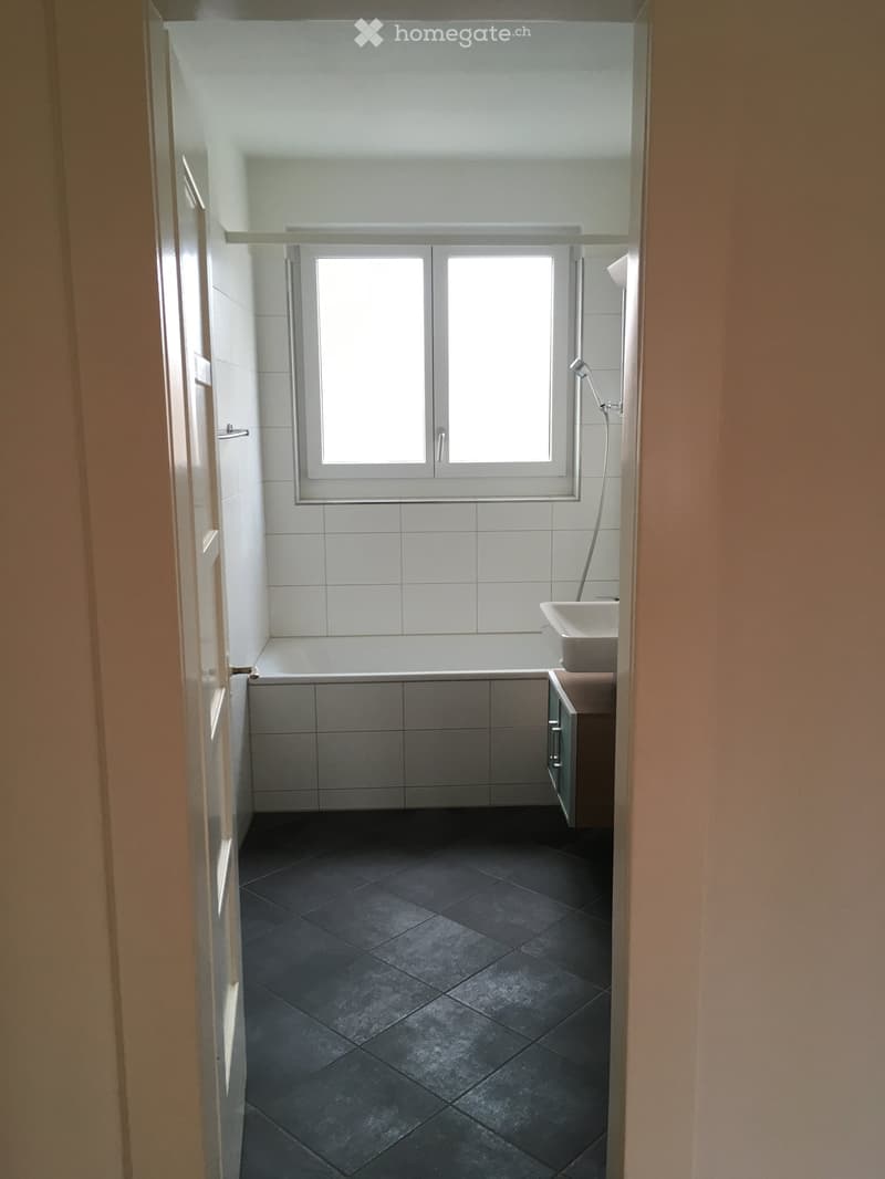 Modern bathroom with dark tiled floor, white tiled walls, bathtub, sink, and window above the tub.