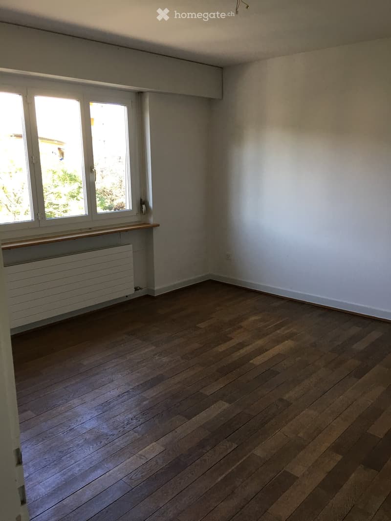 Empty room with wooden flooring, white walls, and large windows with white frames. A radiator is visible beneath the windows.