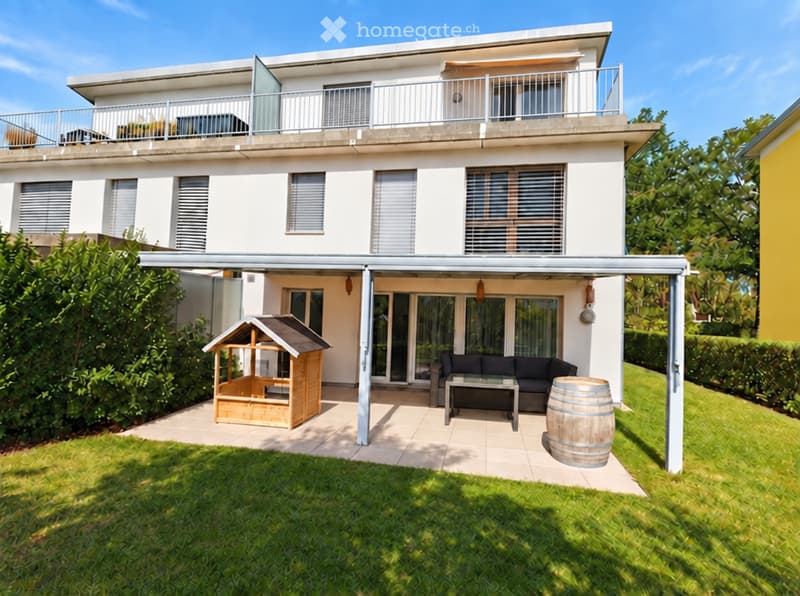 Modern two-story house with white exterior, large windows, covered patio area, and outdoor seating including a sofa, coffee table, and wooden playhouse.