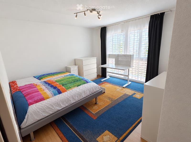 Bright bedroom with a colorful quilted bed, a modern white desk and chair, a dresser, and a nightstand. Large windows with black curtains and a blue carpet add to the contemporary feel.