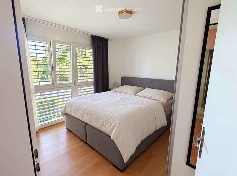 Modern bedroom with a large bed, white bedding, grey headboard, wooden flooring, and large windows with blinds and curtains.