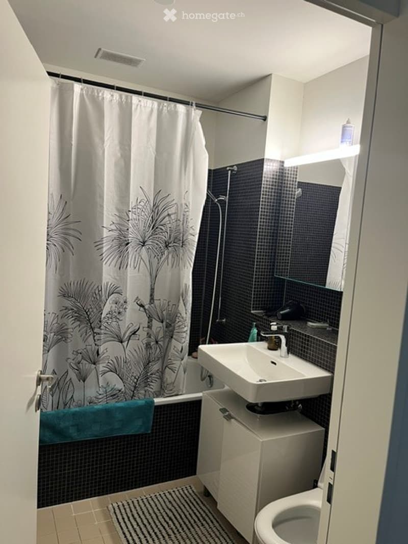 Modern bathroom with white walls, black tiled shower area, a white sink, and a toilet. A decorative shower curtain with a leaf pattern is visible.