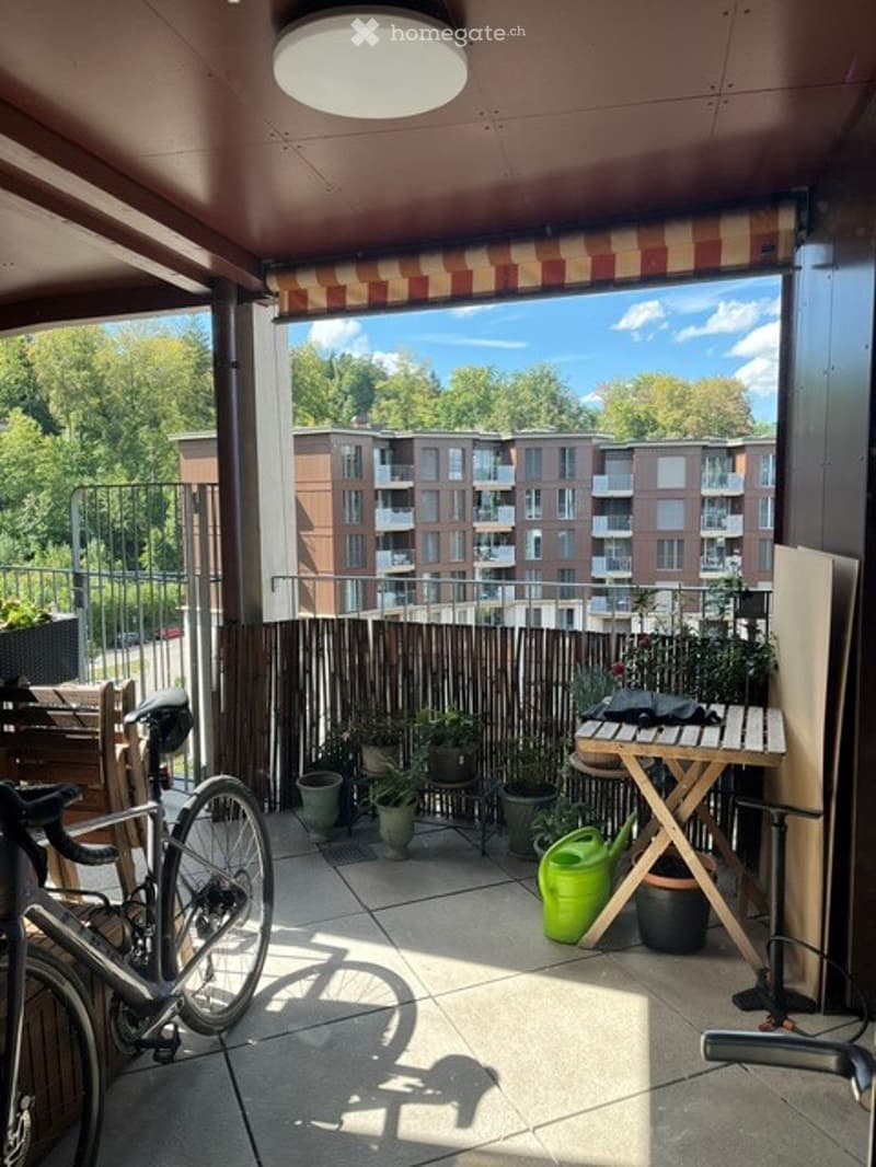 Balcony with outdoor furniture, plants, and a view of a building and greenery.