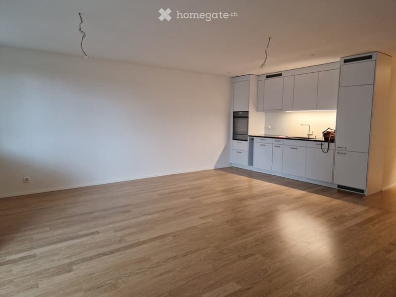 Empty room with light wooden flooring and white walls, featuring a modern kitchen area with cabinets and appliances.