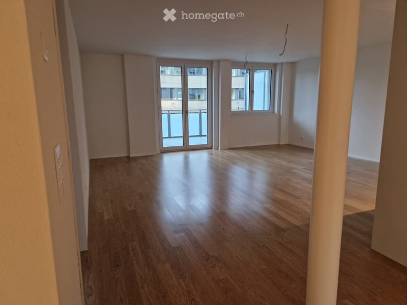 Empty room with wooden flooring, white walls, and large windows with a sliding glass door leading to a balcony.